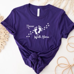 Może przedstawiać: Fioletowy T-shirt z białym napisem "Never Walk Alone" i grafiką dw&oacute;ch ślad&oacute;w st&oacute;p z odciskami łap, kt&oacute;re za nimi podążają.
