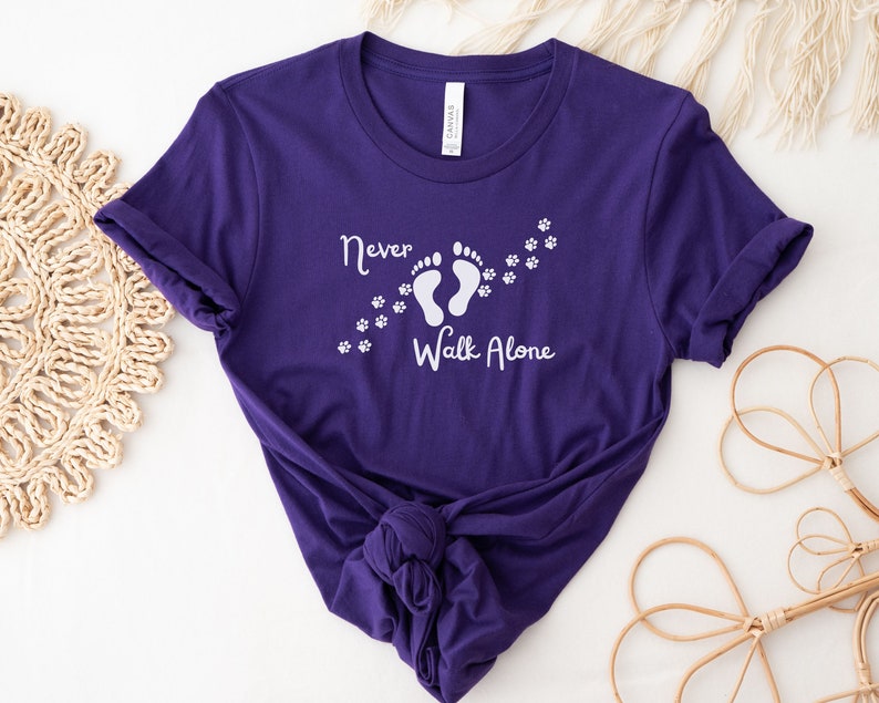 Może przedstawiać: Fioletowy T-shirt z białym napisem "Never Walk Alone" i grafiką dw&oacute;ch ślad&oacute;w st&oacute;p z odciskami łap, kt&oacute;re za nimi podążają.