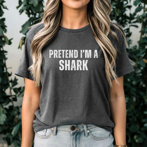May include: Dark grey t-shirt featuring the white text "PRETEND I'M A SHARK". The shirt has a crew neck and is worn by a person. The background is a blurred green foliage.