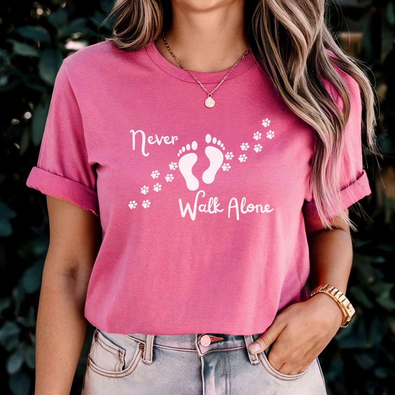 Może przedstawiać: R&oacute;żowa koszulka z białym napisem "Never Walk Alone" i grafiką dw&oacute;ch ślad&oacute;w st&oacute;p z odciskami łap wok&oacute;ł.