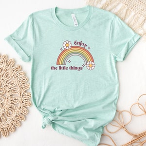 May include: A light blue t-shirt with a rainbow and daisies graphic. The text "Enjoy the little things" is printed on the shirt.