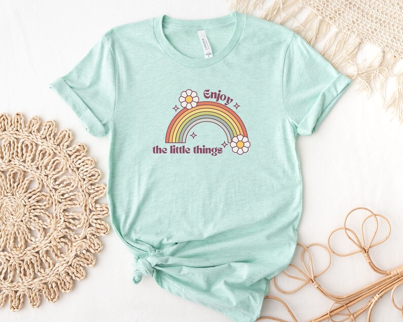 May include: A light blue t-shirt with a rainbow and daisies graphic. The text "Enjoy the little things" is printed on the shirt.