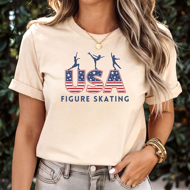 May include: A beige t-shirt featuring the text "USA FIGURE SKATING" in red and blue, with a design of skaters. The word "USA" is stylized with the American flag design. The shirt has short sleeves.