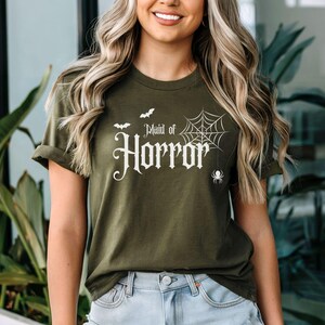 May include: Olive green t-shirt with the text "Maid of Horror" in a gothic font, accompanied by a spider web, bats, and a spider graphic. The shirt is a short-sleeved crew neck.