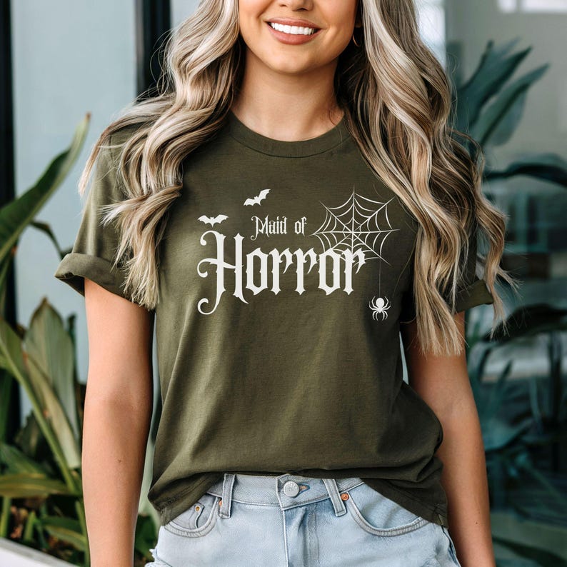 May include: Olive green t-shirt with the text "Maid of Horror" in a gothic font, accompanied by a spider web, bats, and a spider graphic. The shirt is a short-sleeved crew neck.