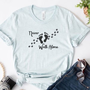 Może przedstawiać: Jasnoniebieska koszulka z napisem "Never Walk Alone" i grafiką dw&oacute;ch ślad&oacute;w st&oacute;p z odciskami łap, kt&oacute;re je śledzą.