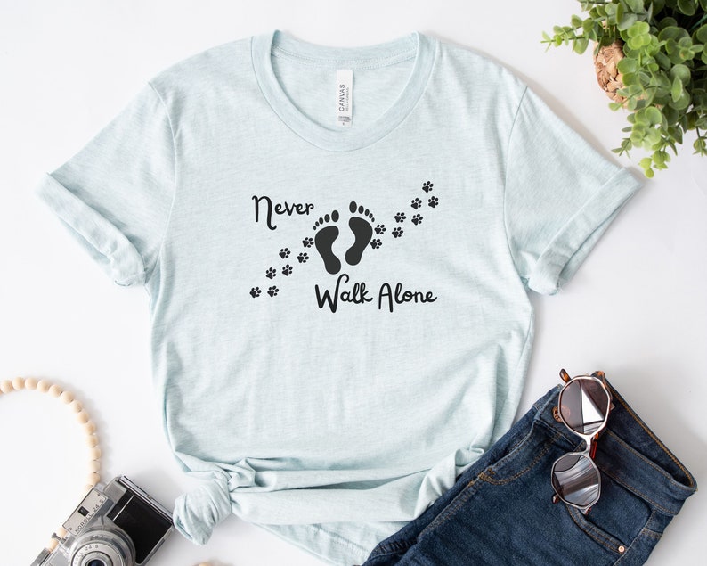 Może przedstawiać: Jasnoniebieska koszulka z napisem "Never Walk Alone" i grafiką dw&oacute;ch ślad&oacute;w st&oacute;p z odciskami łap, kt&oacute;re je śledzą.