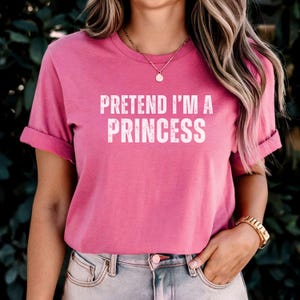 May include: A pink t-shirt with the white text "PRETEND I'M A PRINCESS". The shirt has rolled sleeves and is worn with blue jeans. A gold watch and necklace are also visible. The image is set against a green background.