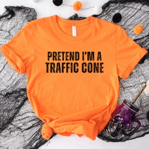 May include: Vibrant orange t-shirt featuring the phrase "PRETEND I'M A TRAFFIC CONE" in bold black text. The shirt is knotted at the hem and lies on black fabric. A purple skull-shaped glass with a straw is visible in the image.