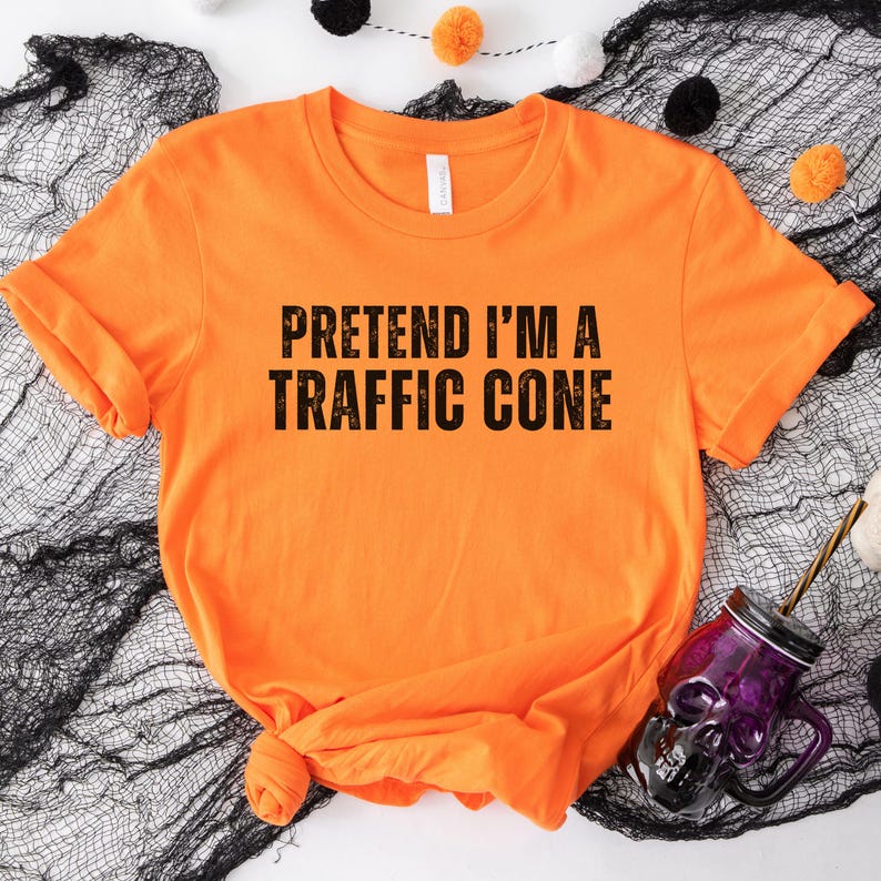 May include: Vibrant orange t-shirt featuring the phrase "PRETEND I'M A TRAFFIC CONE" in bold black text. The shirt is knotted at the hem and lies on black fabric. A purple skull-shaped glass with a straw is visible in the image.