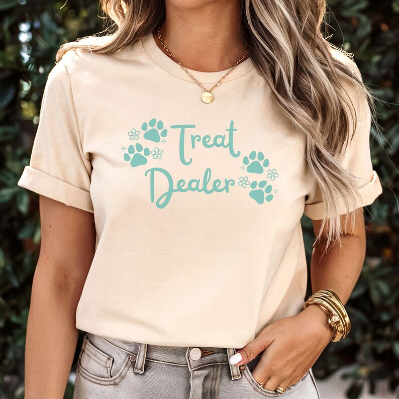 May include: A beige t-shirt with the text "Treat Dealer" in teal blue lettering. The text is surrounded by teal blue paw prints and small white flowers.