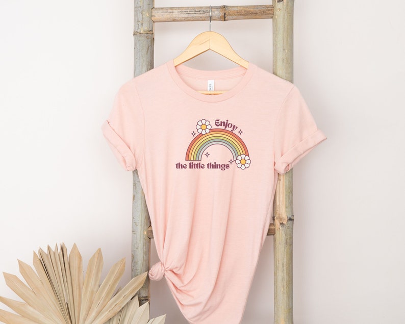 May include: A pink t-shirt with a rainbow and the text "Enjoy the little things" printed on the front. The rainbow is in shades of orange, yellow, green, blue, and purple. There are two white daisies on either side of the rainbow.