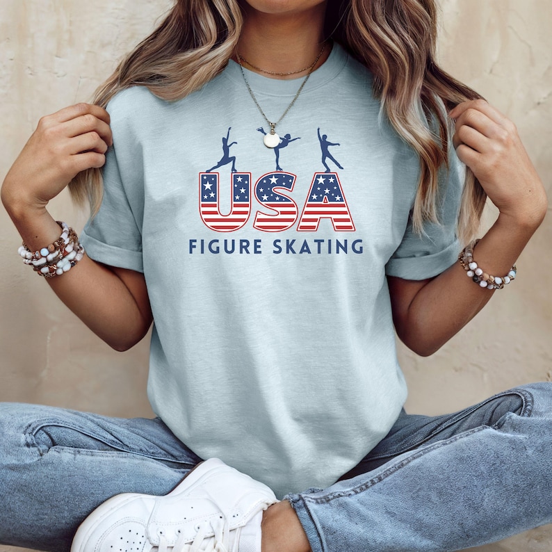 May include: Light blue t-shirt with the text "USA FIGURE SKATING" in red and blue. The word "USA" has a stars and stripes design. Above the text are three silhouettes of figure skaters.