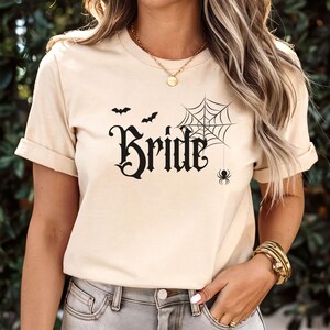 May include: Beige t-shirt with the word "Bride" in a gothic font, accompanied by a spider web, spider, and bats. The shirt has short sleeves and a round neckline. The overall theme is Halloween or a spooky wedding.