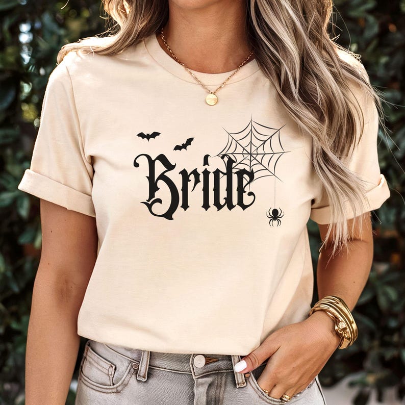 May include: Beige t-shirt with the word "Bride" in a gothic font, accompanied by a spider web, spider, and bats. The shirt has short sleeves and a round neckline. The overall theme is Halloween or a spooky wedding.