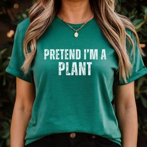 May include: A teal t-shirt featuring the white text "PRETEND I'M A PLANT". The shirt has a round neck and short sleeves. A gold necklace with a pendant is worn.