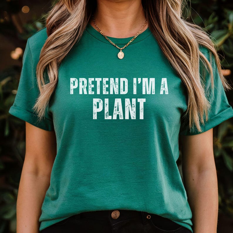 May include: A teal t-shirt featuring the white text "PRETEND I'M A PLANT". The shirt has a round neck and short sleeves. A gold necklace with a pendant is worn.