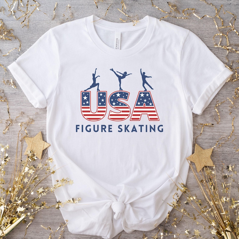 A white t-shirt with a blue and red USA graphic with three figure skater silhouettes on top of the USA letters. The text ‘Figure Skating' is below the graphic.
