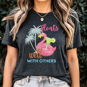 Op de afbeelding: Een donkergrijs T-shirt met een roze flamingo opblaasbaar zwembad drijvend met een margarita glas. De tekst "floats well with others" is in blauw onder de flamingo gedrukt.