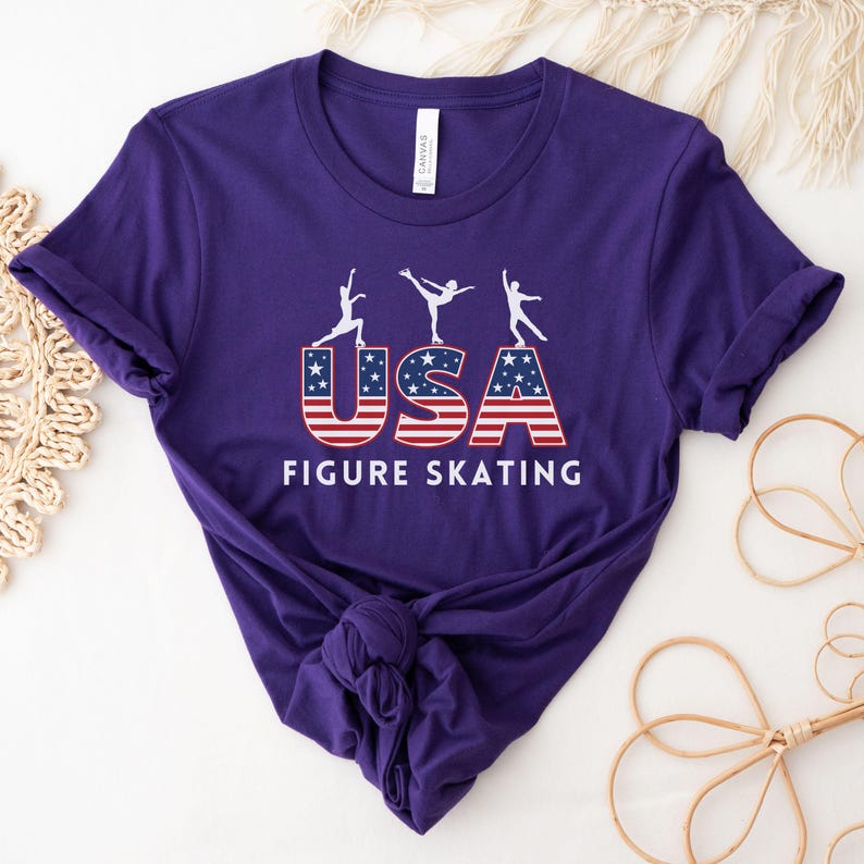 May include: A purple t-shirt featuring the text "USA FIGURE SKATING" in white. The letters "USA" are designed with a red and white striped pattern and blue stars. Above the text are three white silhouettes of figure skaters.