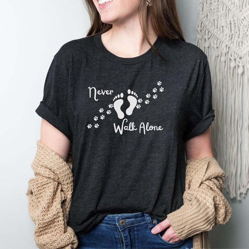 A woman wearing a dark gray heather t-shirt featuring a design with two footprints and paw prints, accompanied by the text 'Never Walk Alone.' The shirt is shown on a model, and appears to be made of a soft, comfortable material.  The design is simple yet meaningful, suitable for pet lovers or those who value companionship.