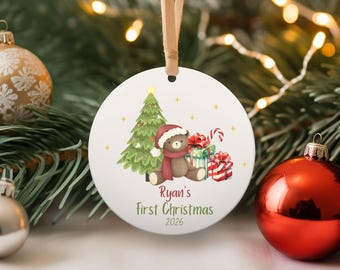 May include: A round white ceramic ornament hanging from a tan ribbon. The design features a brown teddy bear wearing a red hat and scarf, a green Christmas tree, and wrapped gifts. The text reads Ryan's First Christmas 2026 in red and green.