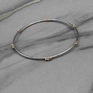 May include: A silver bangle bracelet featuring small gold-coloured bead clusters spaced around the band. The bracelet is presented on a grey and white marble-effect surface.