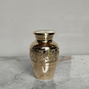 May include: A small, gold-colored urn with a lid. The urn has a rounded body and a decorative band of etched floral patterns around the middle. The surface is shiny and reflective, with a smooth finish. The urn is likely made of metal.