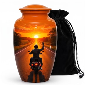 May include: A cylindrical orange cremation urn featuring a printed graphic of a motorcyclist riding into a sunset on a long road, with one hand raised. The urn has a matching orange lid and is accompanied by a black velvet drawstring bag.