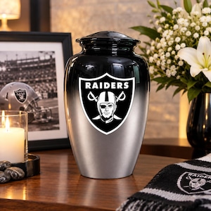 May include: A black and silver metal cremation urn featuring the Raiders team logo on the front. The urn has a smooth, tapered body with a gradient finish, transitioning from a dark black top to a metallic silver base, topped with a lid.