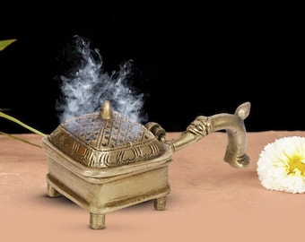 May include: A brass incense burner with a square base and a handle. The burner has a decorative design and is emitting smoke.