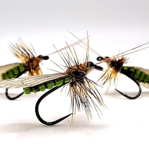 May include: Three artificial fishing flies. Each fly has a black hook, a body with green and brown accents, and translucent wings. The flies are arranged against a white background.