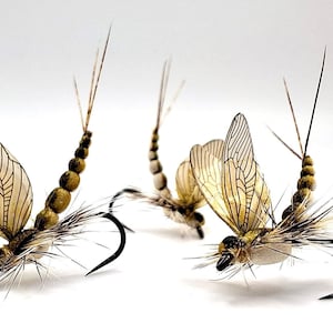 May include: Close-up of three artificial fishing flies. Each fly features tan, veined wings, a segmented body, and a curved black hook. The flies are designed to mimic insects for fly fishing.