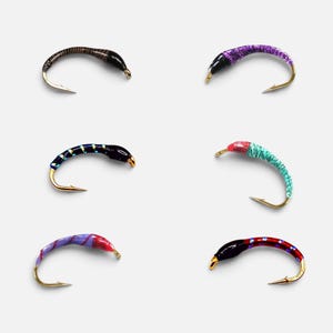 May include: Six curved fishing fly lures with gold-toned hooks. The lures feature various patterns and colours, including brown stripes, purple glitter, black with yellow bands, teal, pink with purple stripes, and red with black segments and blue dots.