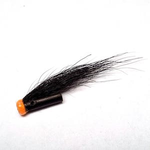 May include: A fishing fly lure featuring a black cylindrical body with a bright orange head and a long, flowing tuft of black hair extending from the top. The lure has a small hole visible on the side of the black tube, designed for line attachment.