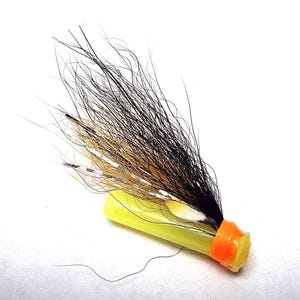 May include: A fishing fly lure featuring a bright yellow plastic tube body with an orange band at the base. The lure is adorned with long, wispy black and tan fibres, interspersed with thin, reflective silver tinsel strands extending outwards.