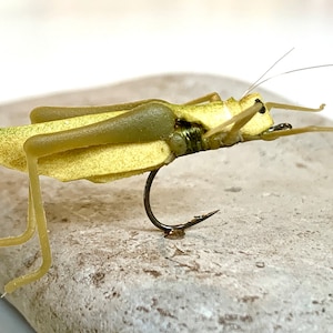 Pode incluir: Um isco de pesca com mosca de cor amarela e verde com forma de gafanhoto realista com um anzuelo preto.