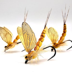Puede incluir: Primer plano de tres moscas de pesca artificiales. Las moscas tienen cuerpos amarillos, alas color canela con detalles de venas negras y anzuelos negros. Las alas están extendidas, sobre un fondo blanco.