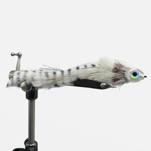 May include: A handmade fly fishing lure with a white and gray speckled body, iridescent accents, and a white head with a detailed eye. The lure is mounted on a silver metal stand, showcasing its intricate design.