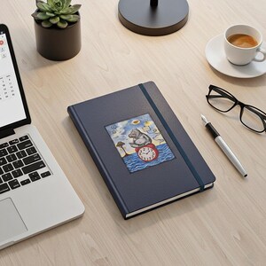 Puede incluir: Un diario azul marino con cierre de banda elástica. La portada presenta una colorida ilustración de un hipopótamo sentado en un reloj. Una computadora portátil, un bolígrafo, gafas y una taza de café también están en el escritorio de madera.