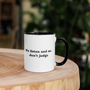 Puede incluir: Taza de cerámica blanca con interior y asa negros. La taza presenta el texto "We listen and we don't judge" en negro. La taza está sobre una superficie de madera.