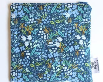 May include: A square zippered pouch featuring a dark blue fabric base with a dense botanical print of light blue, white, and green flowers and foliage. A small white fabric tag on the side displays the text Magic Bag Co. Made in the USA.