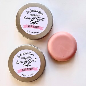 May include: Two round tins with gold lids and white labels that read "So Excitable Soaps Handcrafted Love At First Sight Solid Lotion 2.5 oz / 71 grams". One tin is open and a round, pink solid lotion bar is next to it.
