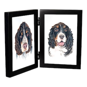 May include: A black double picture frame with two watercolor paintings of Springer Spaniel dogs. The dogs are both looking at the camera. The frame is closed.