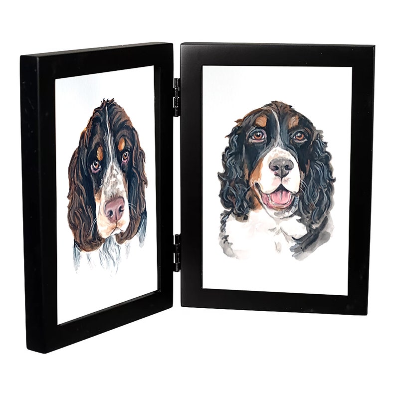 May include: A black double picture frame with two watercolor paintings of Springer Spaniel dogs. The dogs are both looking at the camera. The frame is closed.
