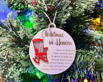 Puede incluir: Un adorno navideño de madera blanca con una mecedora roja y el texto "Navidad en el cielo". El adorno tiene una cita sobre los seres queridos que han fallecido, sugiriendo que vienen a la tierra para pasar tiempo contigo. Te anima a guardarles un asiento en la mesa, aunque no los veas.