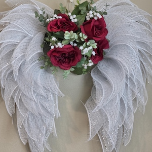 May include: A decorative angel wing wall hanging. The wings are made of white mesh with silver accents. A cluster of red roses, green leaves, and white filler flowers are at the center. The wings are symmetrical and have a feathery appearance.