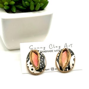 May include: Oval-shaped handmade earrings with a black and white speckled pattern and a gold-colored border. The center of each earring features a pink and peach-colored design. The earrings are displayed on a white card with the text "Sunny Clay Art".