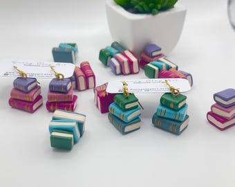 May include: A pair of earrings featuring stacks of miniature books with gold accents. The books are in shades of teal, blue, green, pink, and purple. The earrings are handmade and have gold hooks.