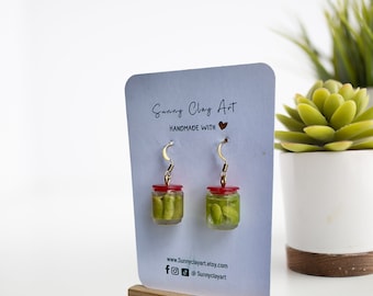 May include: A pair of dangle earrings featuring miniature glass jars filled with green edamame beans. The jars have red lids and gold tops. The earrings are displayed on a white card with the text "Sunny Clay Art Handmade with ♥" and a website address.